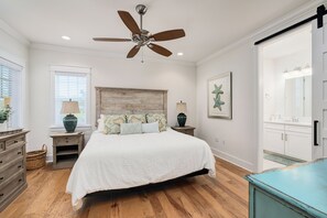 A bedroom with a wooden headboard, a bed with white bedding, a ceiling fan, and a large window with blinds.