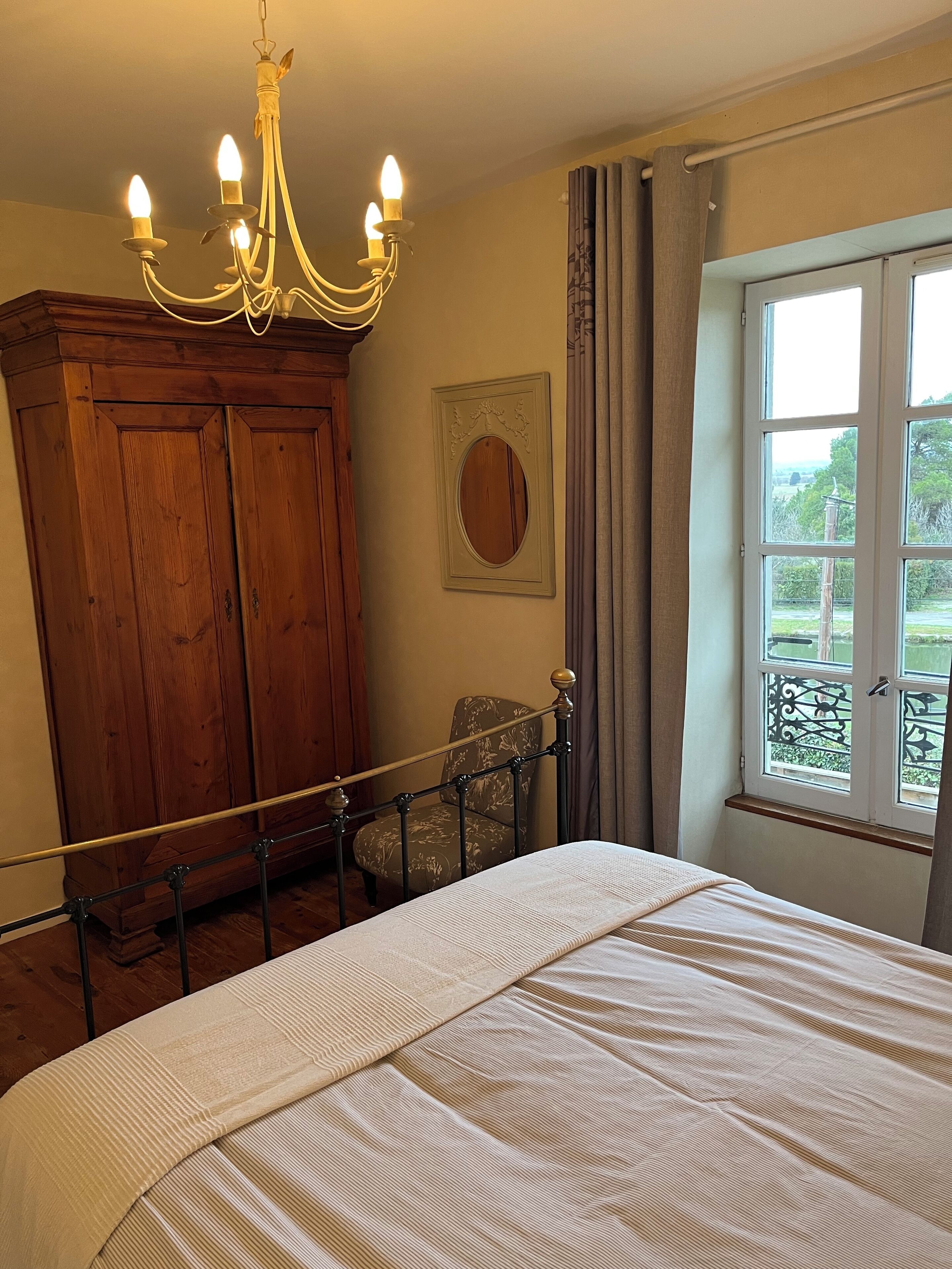 A bedroom with a bed, a wooden wardrobe, a chandelier, a window with a view, and a chair.