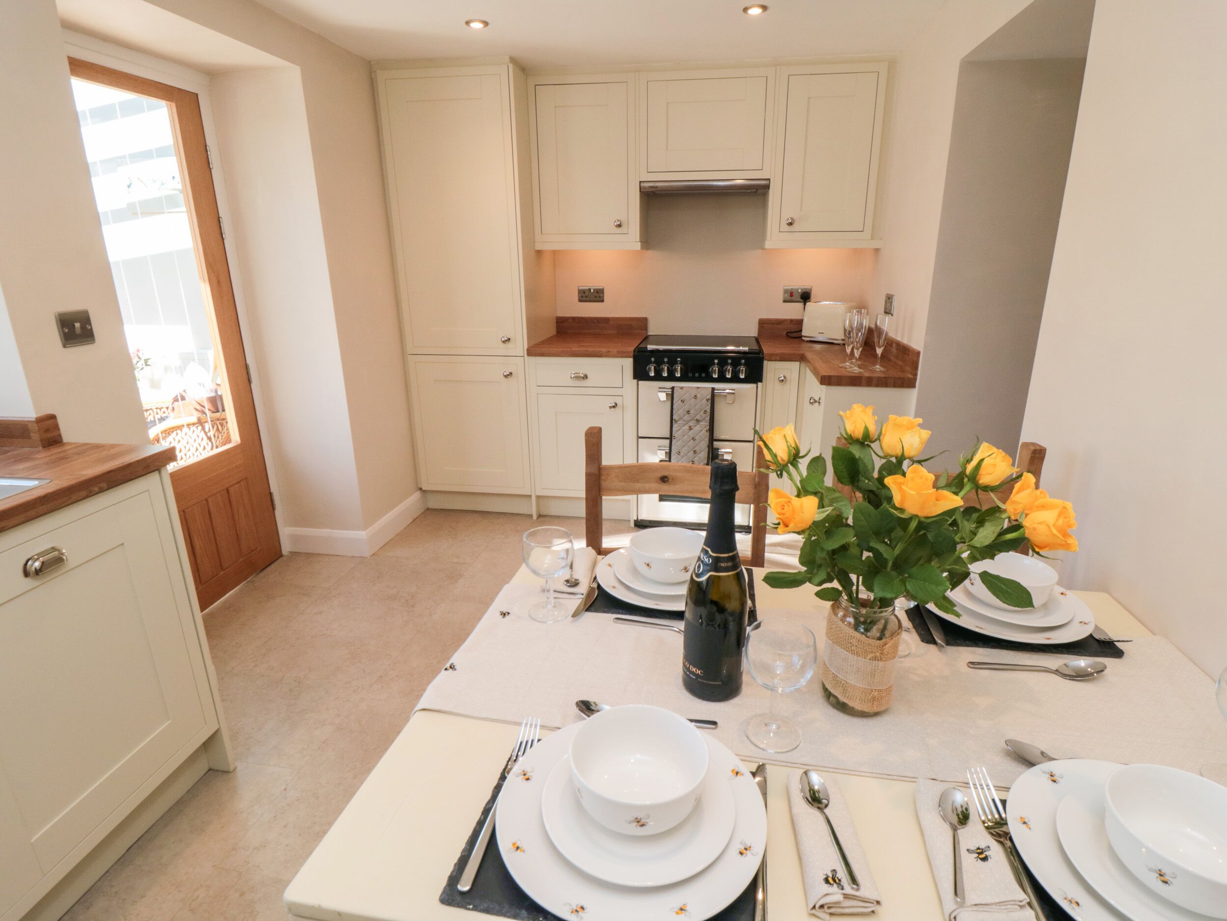 A dining area with a table set for four, a bottle of wine, and a vase of yellow roses.