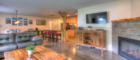 A modern living room with a leather sofa, a wooden coffee table, and a red rug. There is a kitchen area with wooden cabinets and a fireplace.