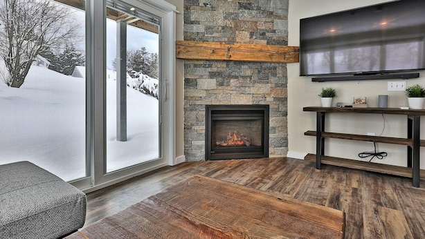 A modern living room with a stone fireplace, a large TV mounted on the wall, and a wooden bench. - Killington Center Inn & Suites by Killington VR - 1 Bedrooms (Killington)