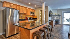 A modern kitchen with wooden cabinets, stainless steel appliances, a large island with stools, and a stone fireplace. - Killington Center Inn & Suites by Killington VR - 1 Bedrooms (Killington)