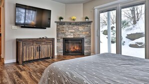 A bedroom with a stone fireplace, a large TV mounted on the wall, and a bed with a patterned bedspread. - Killington Center Inn & Suites by Killington VR - 1 Bedrooms (Killington)