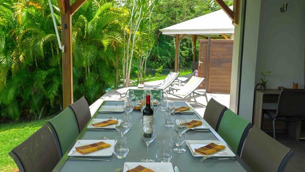 Une table dressée pour un repas, avec une bouteille de vin, des assiettes et des couverts, entourée de chaises, dans un cadre tropical en plein air.