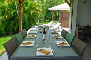 Mesa preparada para una comida con botella de vino, platos y cubiertos, rodeada de sillas, en un entorno tropical al aire libre.