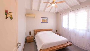 Une chambre avec un lit simple, un ventilateur de plafond, une table de chevet, un climatiseur fixé au mur et une fenêtre avec des rideaux.