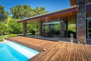 Une maison moderne dotée d’une terrasse en bois, d’une piscine et d’un espace extérieur couvert avec un barbecue.