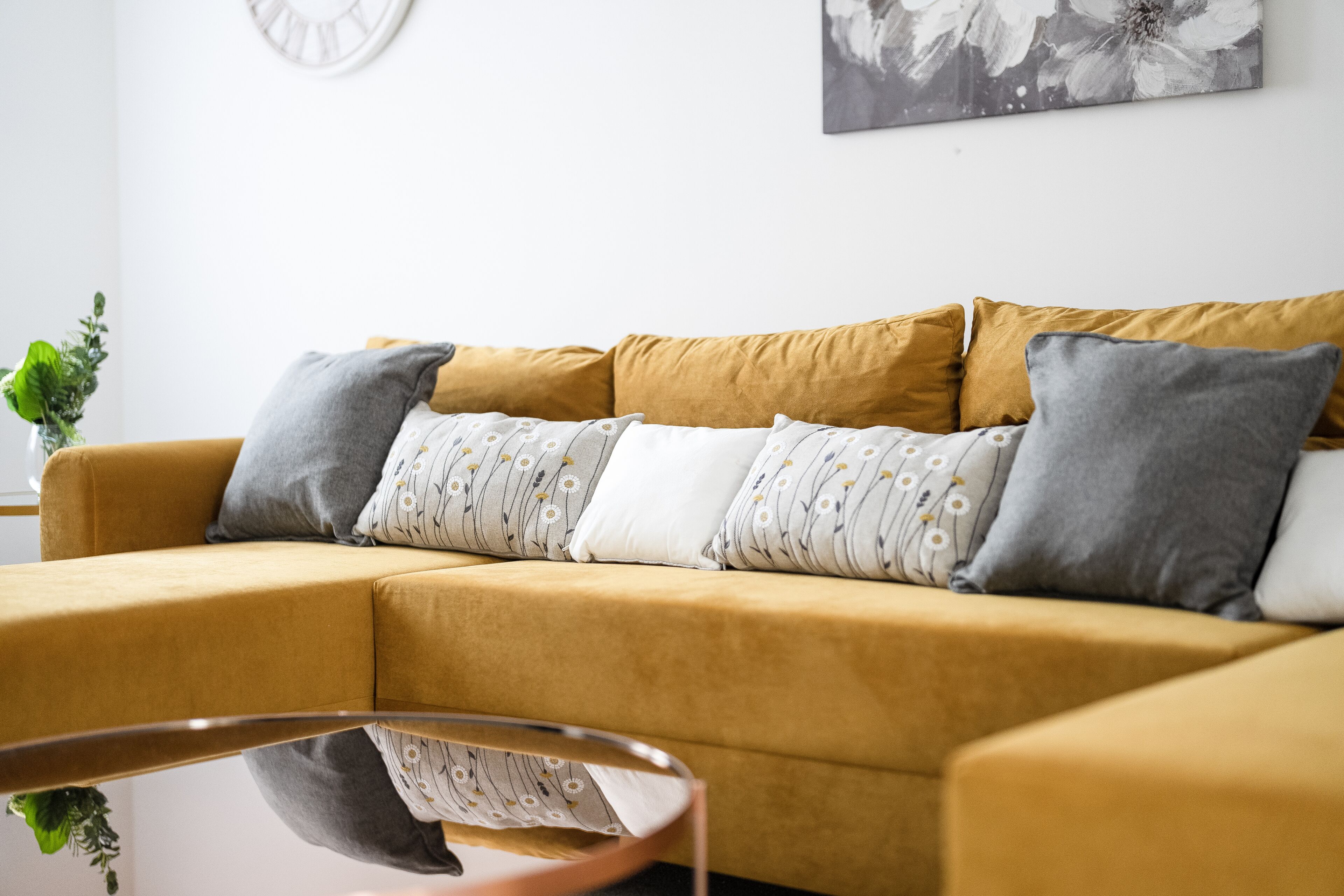 A mustard-colored sofa with patterned pillows, a glass coffee table, and a wall-mounted clock.