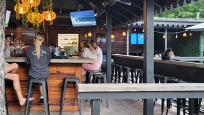 Un espace bar extérieur avec des tables en bois et des tabourets noirs, des lampes suspendues en osier et un écran de télévision fixé au mur.
