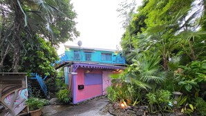 Une maison aux couleurs vives, dotée d’un balcon bleu, entourée d’une végétation luxuriante et bordée d’un chemin en pierre.