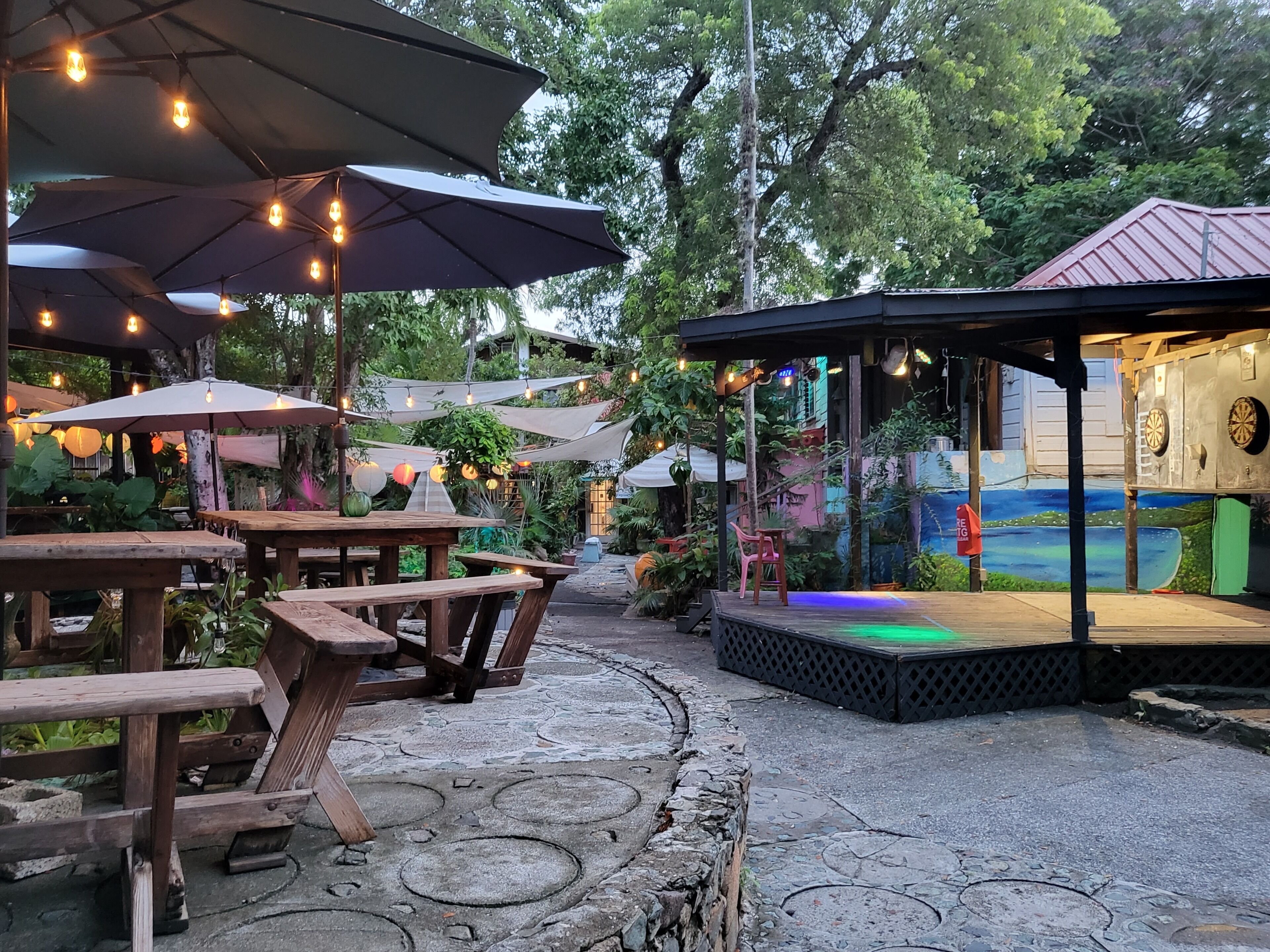 Een terras buiten met houten tafels en parasols, omgeven door groen.
