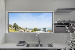 Una cocina con un ventanal que da a un pueblo costero, un lavabo doble y una repisa con tazas y cuencos.