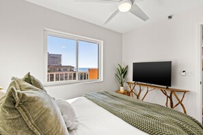 A bedroom with a bed, a television on a wooden stand, a window with a view of buildings, and a ceiling fan. - Beachfront w/ Views! Kid & Pet-friendly, Heated Pool, Spa, Pickle Ball/Tennis (Indian Rocks Beach)