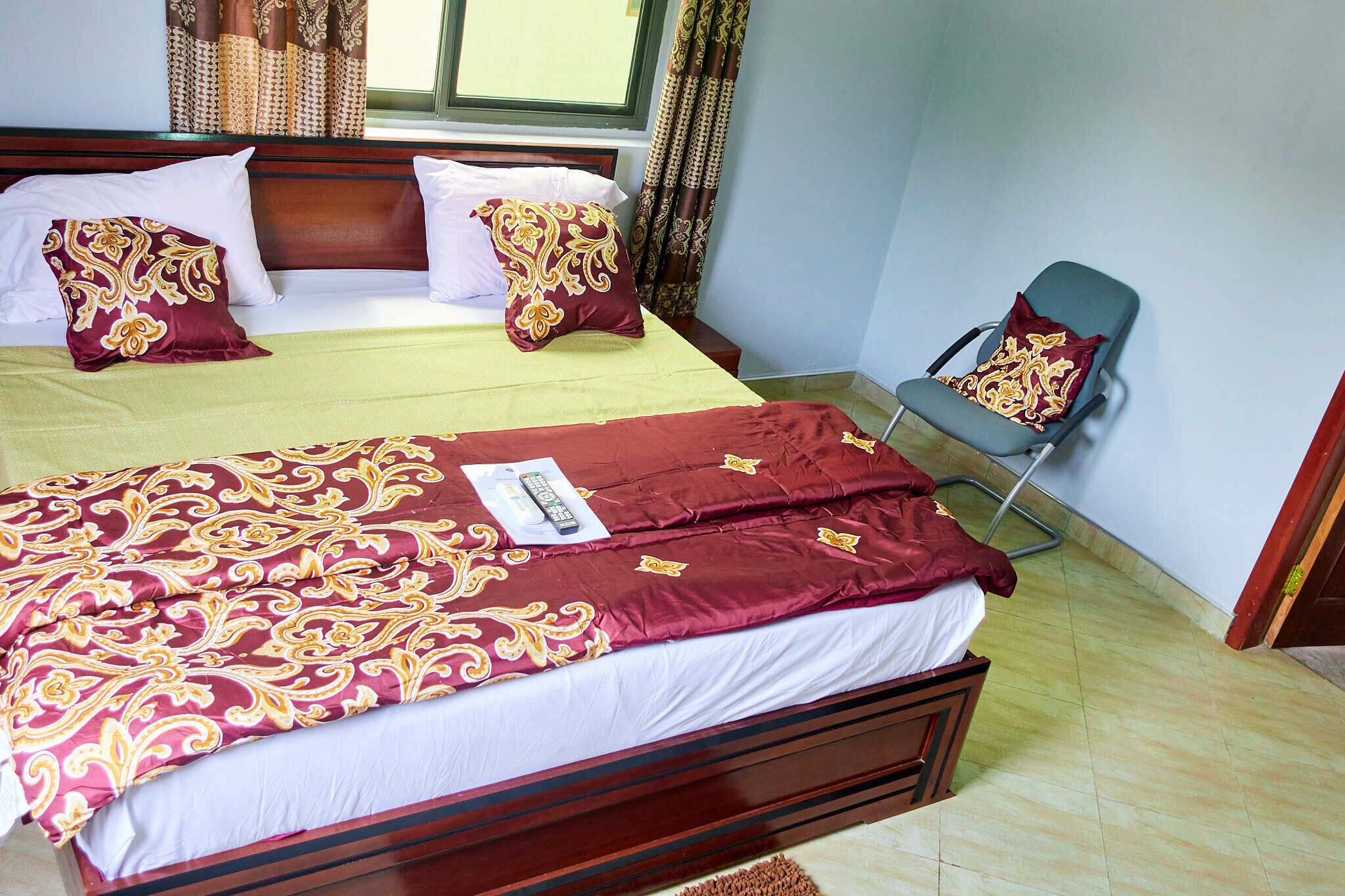 A neatly made bed with a patterned bedspread and pillows, a chair with a cushion, and a window with curtains.