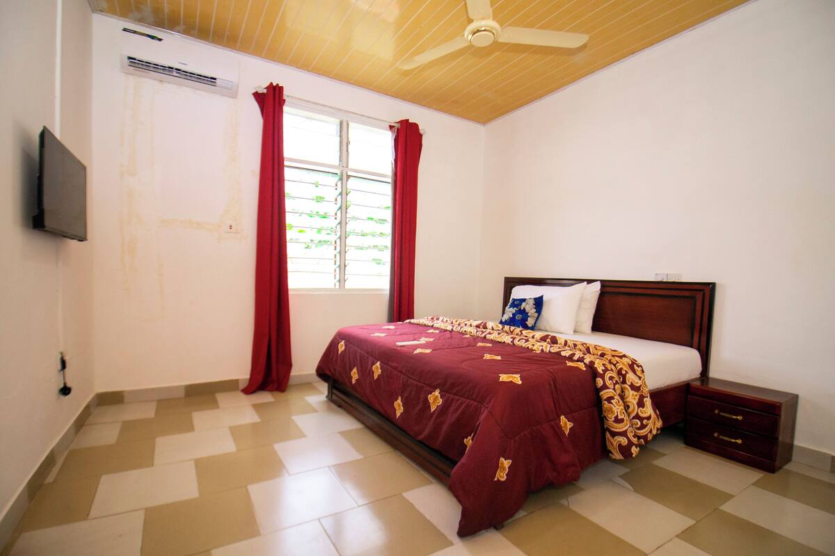 A bedroom with a bed, a nightstand, a TV, and a window with curtains.