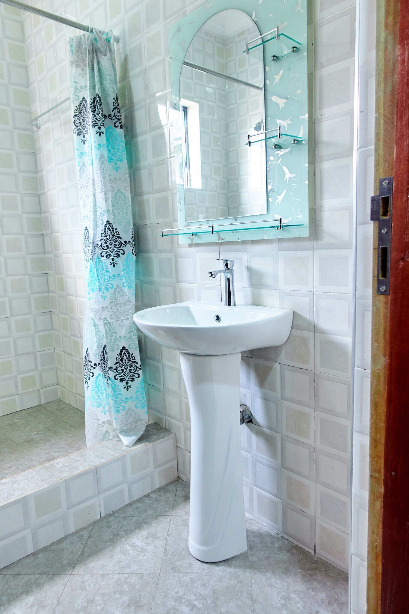 Un baño con ducha de azulejos blancos, lavamanos pedestal blanco, estante de vidrio sobre el lavamanos y cortina de ducha estampada.