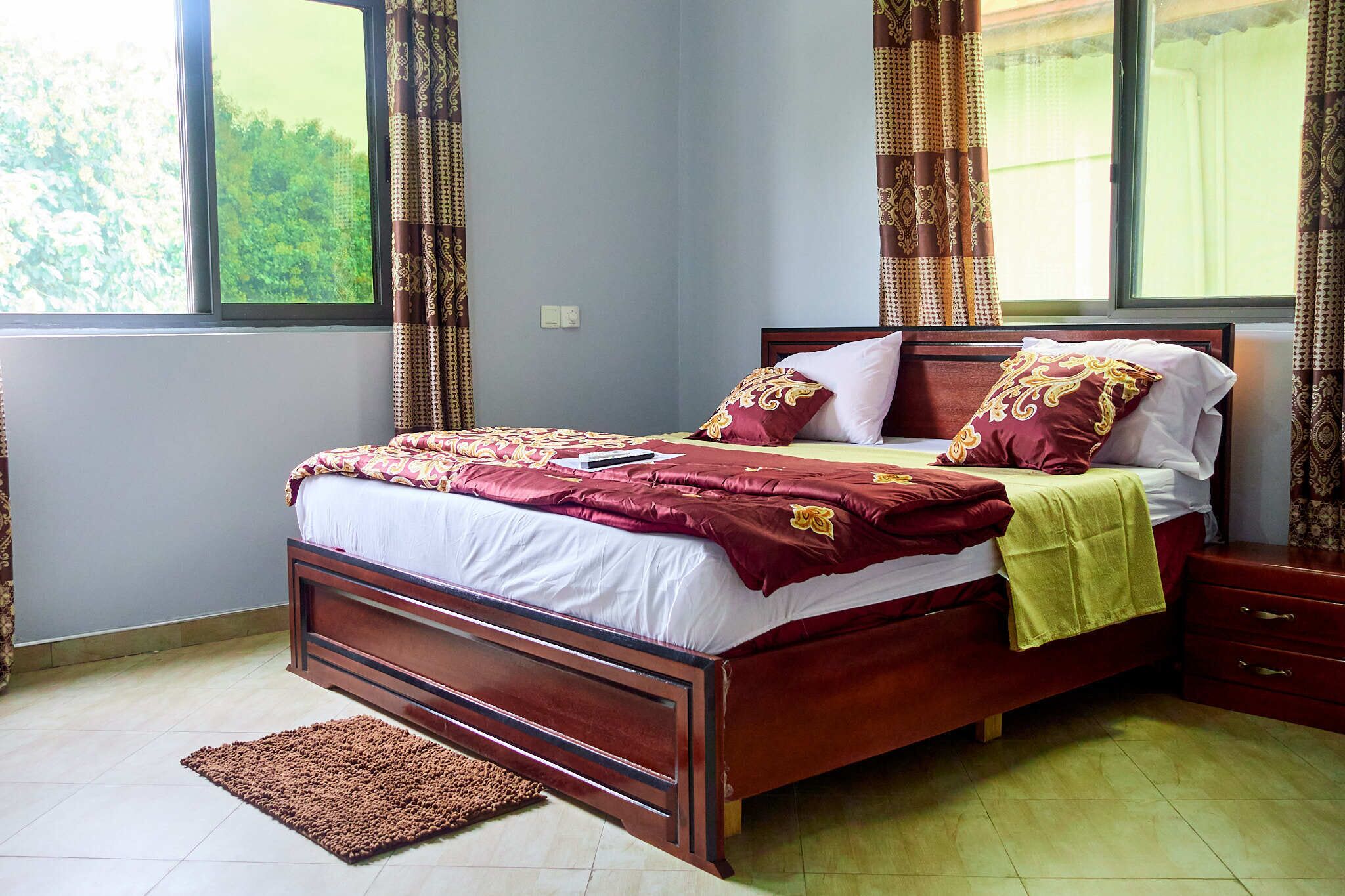 A neatly made bed with a wooden frame, a brown rug, and a window with curtains.
