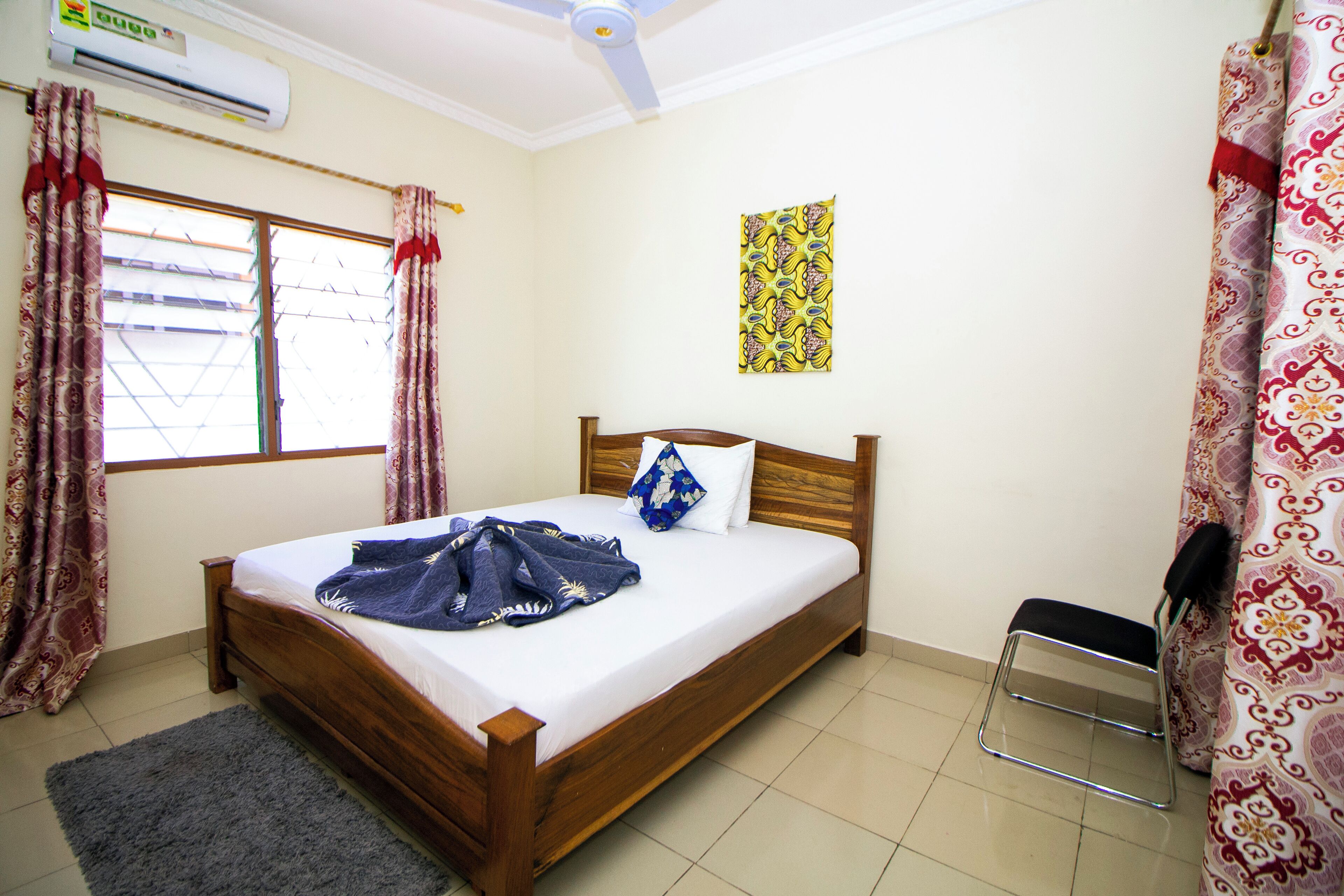 A bedroom with a wooden bed, a ceiling fan, an air conditioner, a window with curtains, a wall hanging, and a chair.