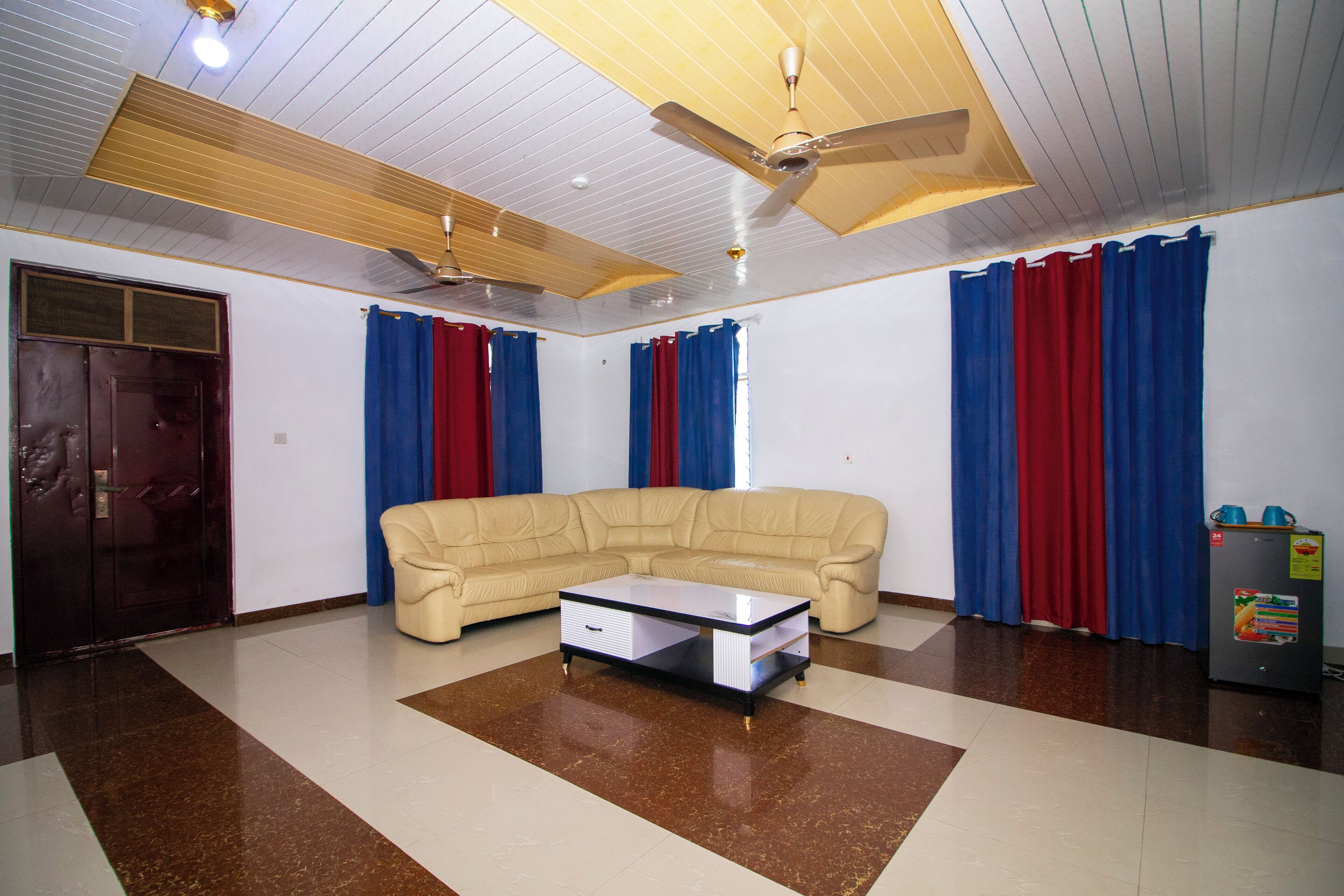 Una sala de estar con un sofá color beige, una mesa de centro de vidrio, un ventilador de techo y cortinas rojas y azules.