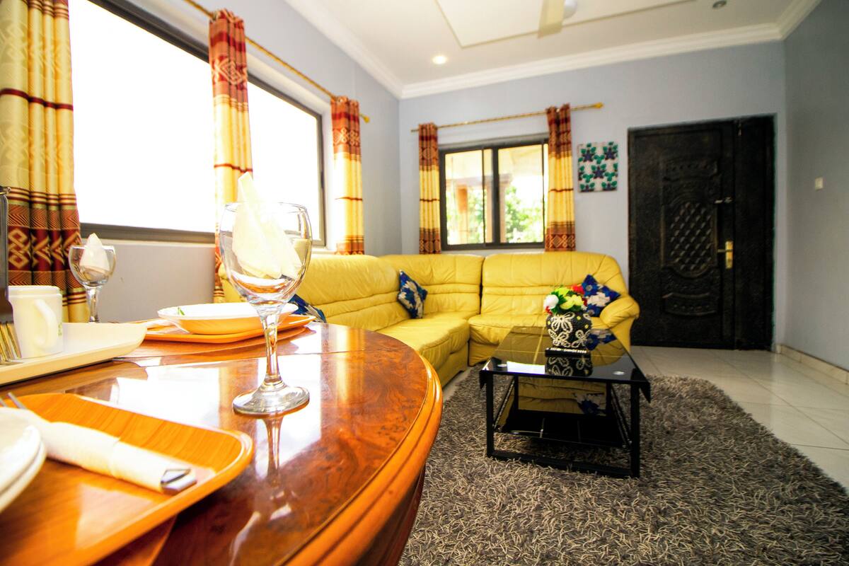 A living room with a yellow sofa, a wooden coffee table, and a dining area with a wooden table and chairs.