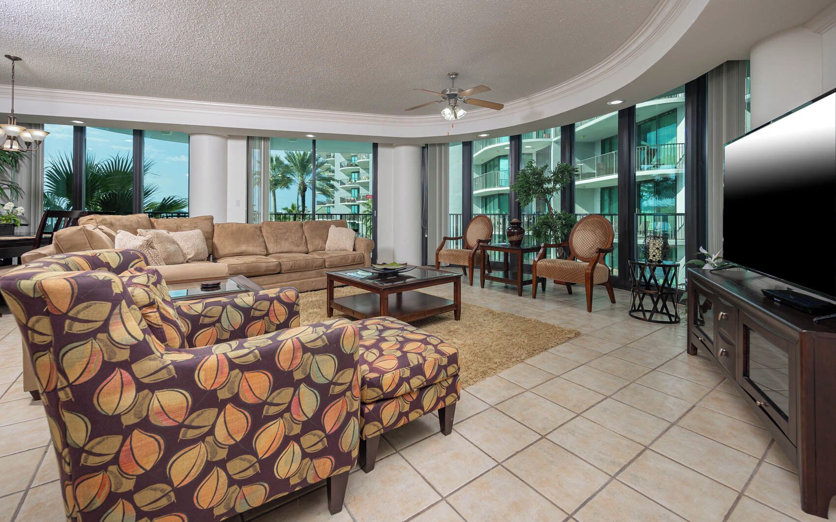 A spacious living room with a large sofa, matching armchairs, a coffee table, and a flat-screen TV. The room features large windows offering views of a building with balconies.