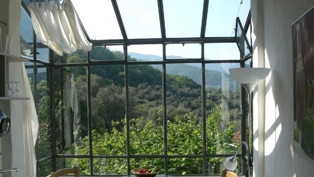 Ein Zimmer mit großem Fenster und Blick auf eine üppige, grüne Landschaft. Es steht ein kleiner runder Tisch mit einer Obstschale, zwei Holzstühle und eine Stehlampe im Raum.