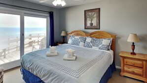 Una camera da letto con un letto, una ventola a soffitto, un comodino, una lampada, un quadro incorniciato e vista sull'oceano tramite una porta scorrevole.