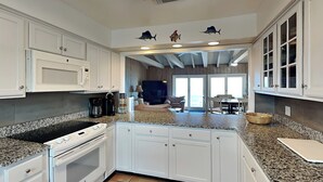 Una cucina moderna con mobili bianchi, piani di lavoro in granito e un'isola centrale. Sono presenti luci a soffitto e si intravede l'area pranzo.