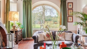 Ein gemütliches Wohnzimmer mit Kamin, einer bequemen Couch und Blick auf den Garten durch ein großes Rundbogenfenster.