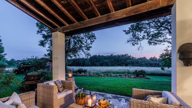 Eine Terrasse im Freien mit Korbmöbeln, einem Tisch mit Obst und Kerzen sowie Blick auf ein grünes Feld und Bäume.