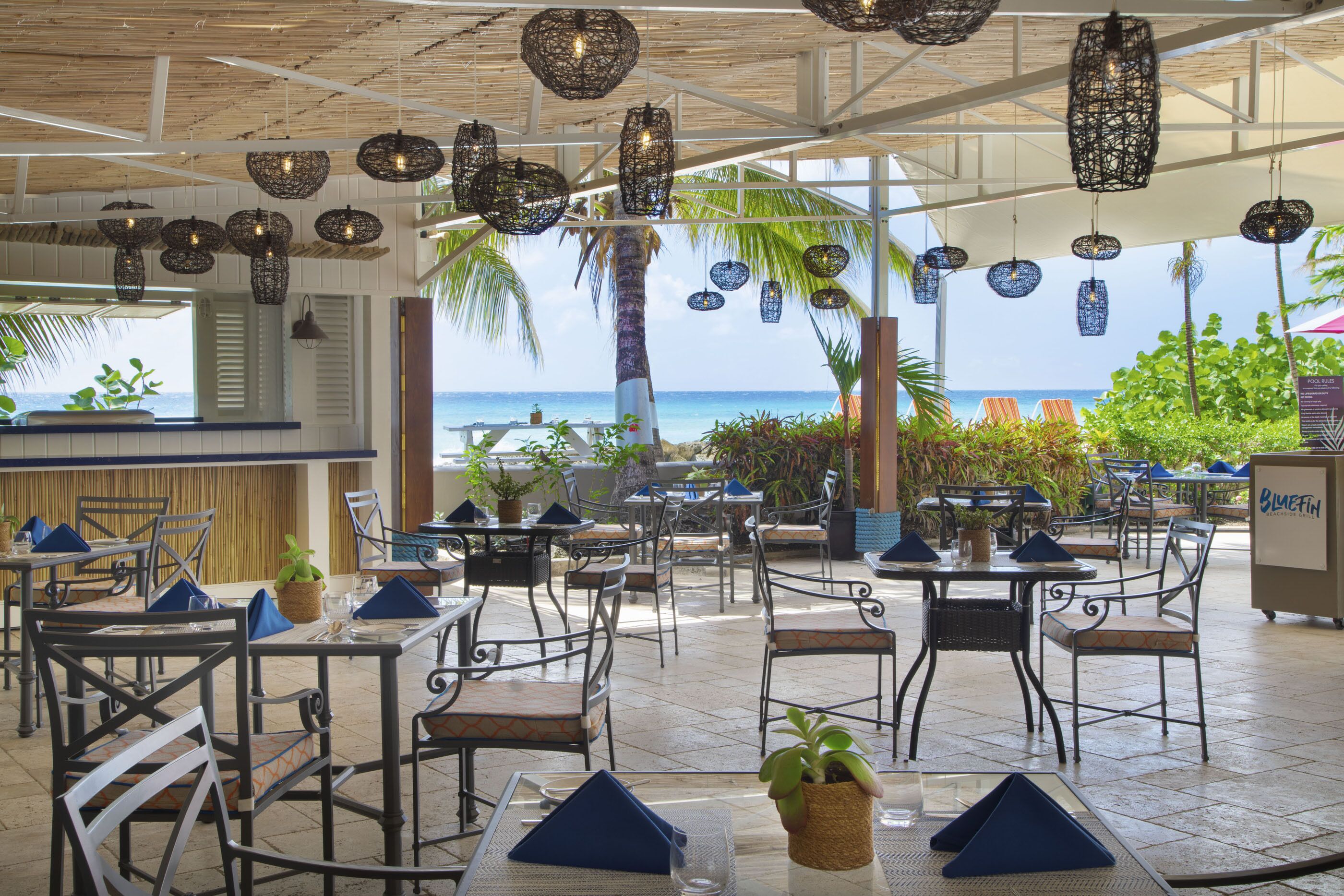 An outdoor dining area with tables and chairs, hanging wicker light fixtures, and a view of the ocean.
