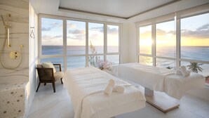A spa room with two beds, a massage table, a chair, and a large window with a view of the ocean. - Concierge Collection at O2 Beach Club & Spa by Ocean Hotels (St. Lawrence Gap)