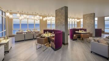 Un comedor moderno con varias mesas y sillas, ventanas amplias con vista al mar e iluminación colgante.