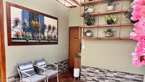 A room with a tiled floor, a chair with cushions, a wall-mounted shelf with potted plants, and a framed picture of a building with flowers. - CASA HIDALGO ECUADOR (Cumbayá)