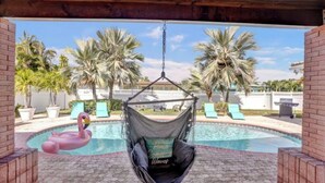 A hanging chair by a pool with a flamingo float, palm trees, and a brick wall.
