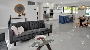 A modern living room with a grey sofa, a glass coffee table, and a kitchen in the background.
