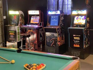 Una mesa de billar con diferentes billas, juegos de arcade y un simulador de baloncesto al fondo.