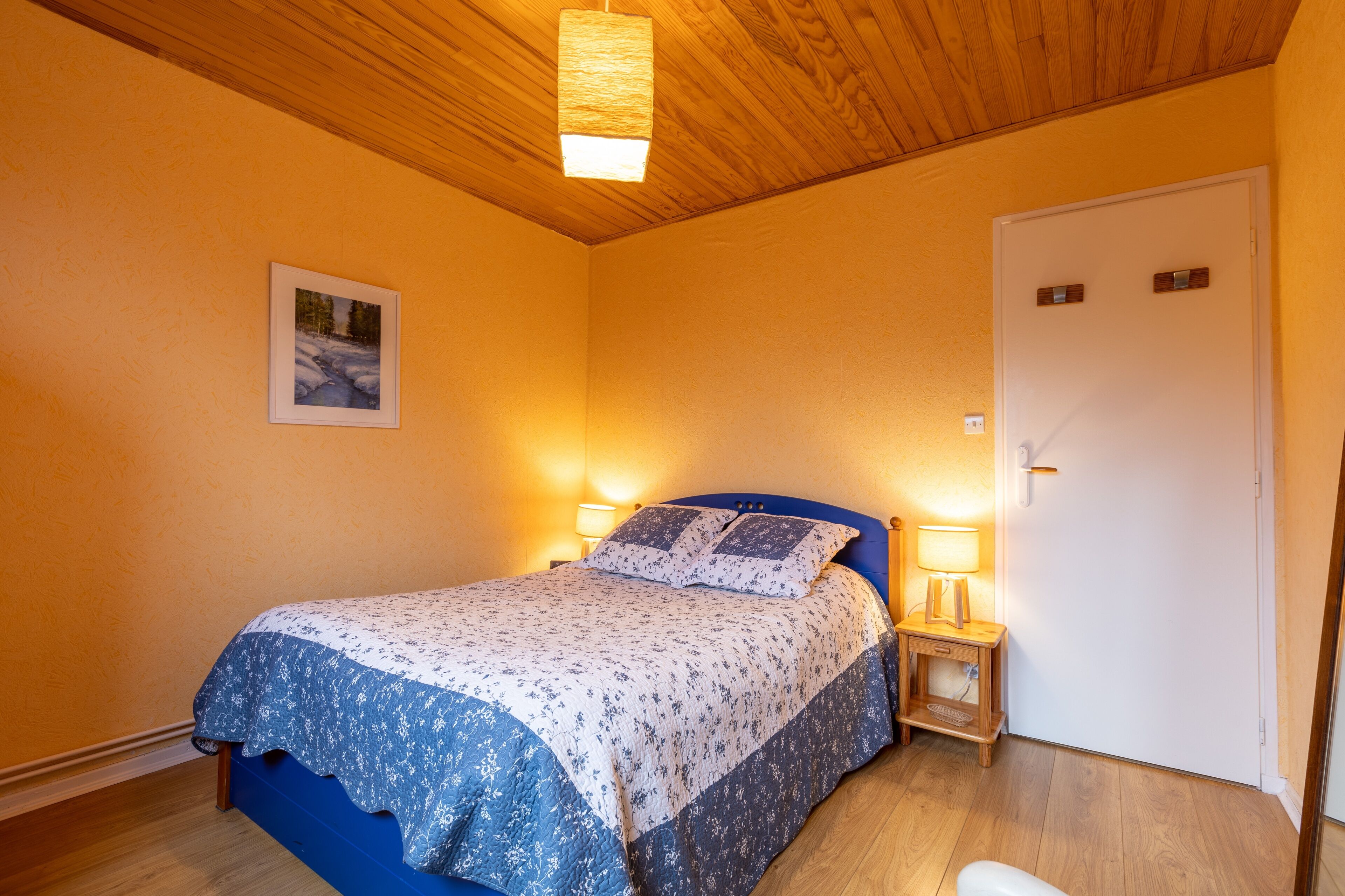 A bedroom with a bed, a bedside table with a lamp, a door, and a framed picture on the wall.