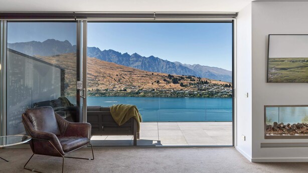 A modern room with a large sliding glass door leading to a balcony with a lake view, a leather armchair, and a framed picture on the wall. - Remarkable Lake View Modern Executive Home (Queenstown)