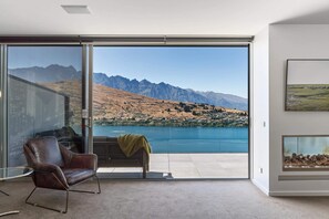 A modern room with a large sliding glass door leading to a balcony with a lake view, a leather armchair, and a framed picture on the wall. - Remarkable Lake View Modern Executive Home (Queenstown)