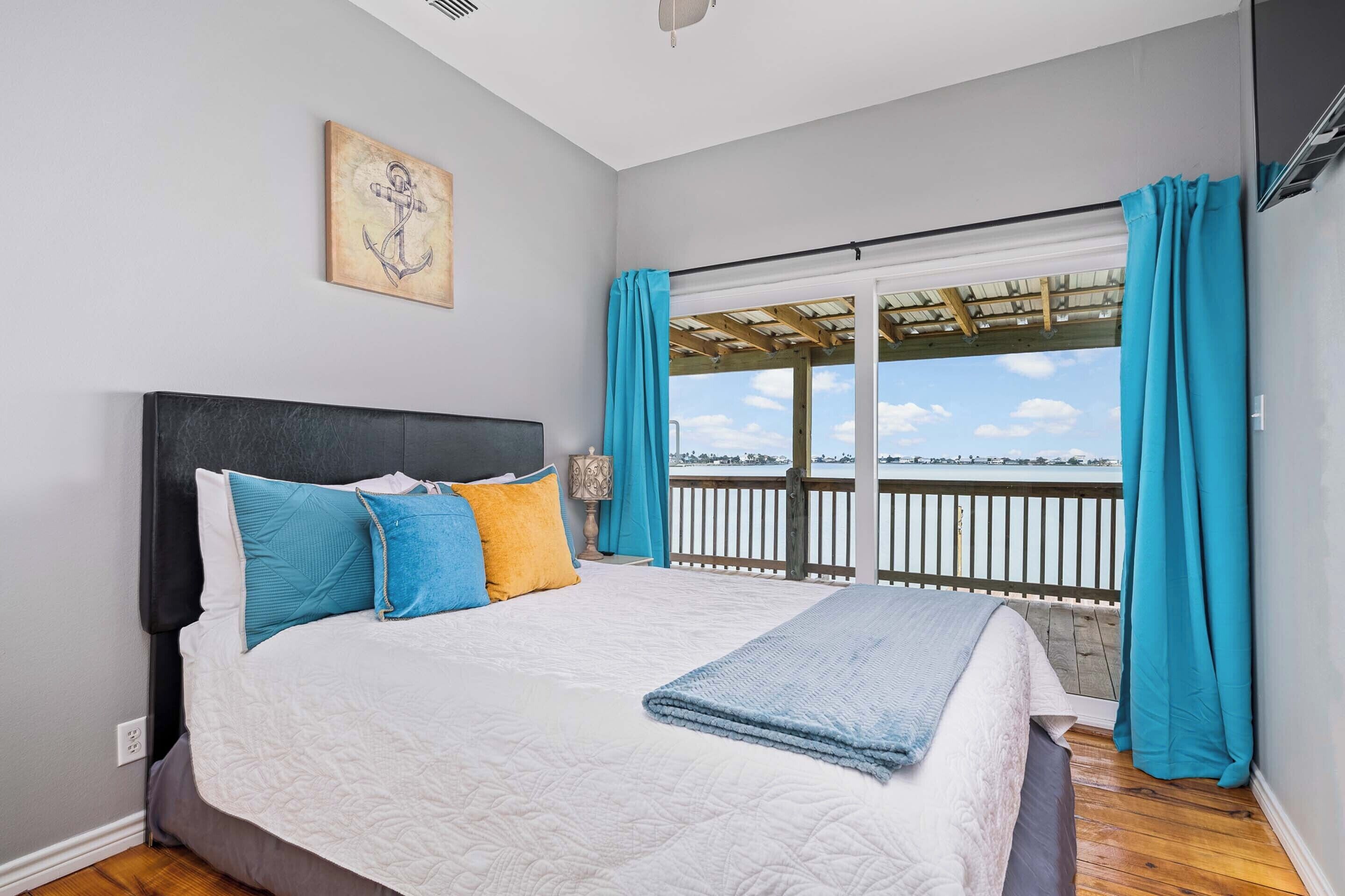 A bedroom with a large bed, a view of the sea, and a wooden deck.