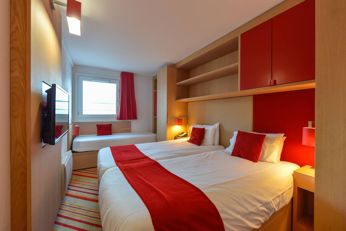 Habitación de hotel con una cama grande, una cama más pequeña, un escritorio, un televisor y una ventana con cortinas rojas.