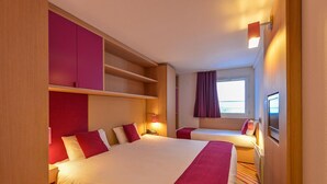 Ein Hotelzimmer mit einem großen Bett, einem kleineren Bett, einem Kleiderschrank, einem Nachttisch mit Lampe und einem Fenster mit Vorhängen.