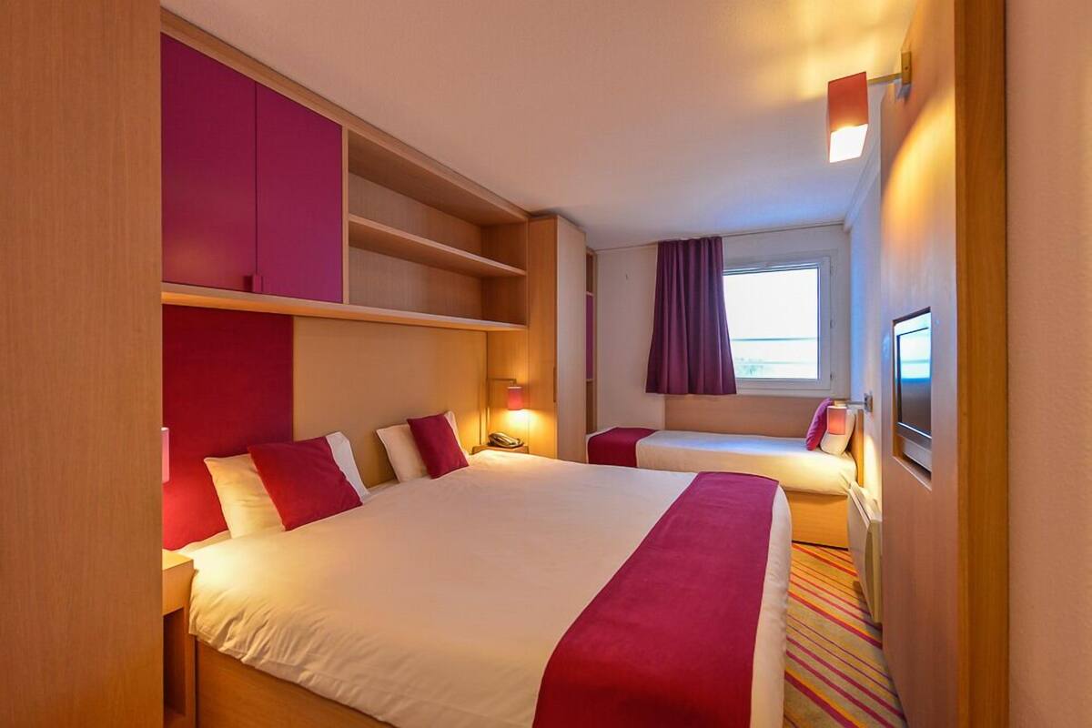 Habitación de hotel con una cama grande, una cama individual, un armario, una mesita de noche con lámpara y una ventana con cortinas.