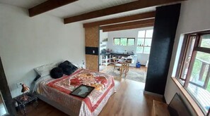 A bedroom with a large bed, a bedside table, and a view of a kitchen and dining area.
