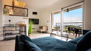 Una sala moderna con un sofá grande, una mesa de centro y un televisor en la pared. Hay un balcón con asientos exteriores y vistas al paisaje.