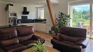 Een moderne keuken met een zwart aanrecht, roestvrijstalen apparatuur en houten balken. Er staan twee leren banken en een houten salontafel met een plant. Een groot raam biedt uitzicht op een groen landschap.