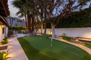 A well-maintained golf course with a putting green, surrounded by trees and a white wall.