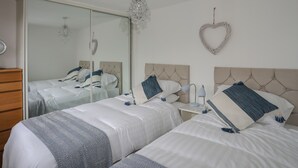 A bedroom with two beds, a dresser, a mirror, a chandelier, and a heart-shaped decoration on the wall. - Bramble Bank (Harlech)