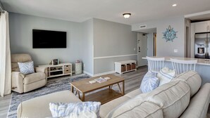 A living room with a beige sofa, a wooden coffee table, a TV mounted on the wall, and a kitchen area with white cabinets and a blue decorative item on the wall. - Sunskipper E2 - 2 bedroom ocean view condo (Carolina Beach)